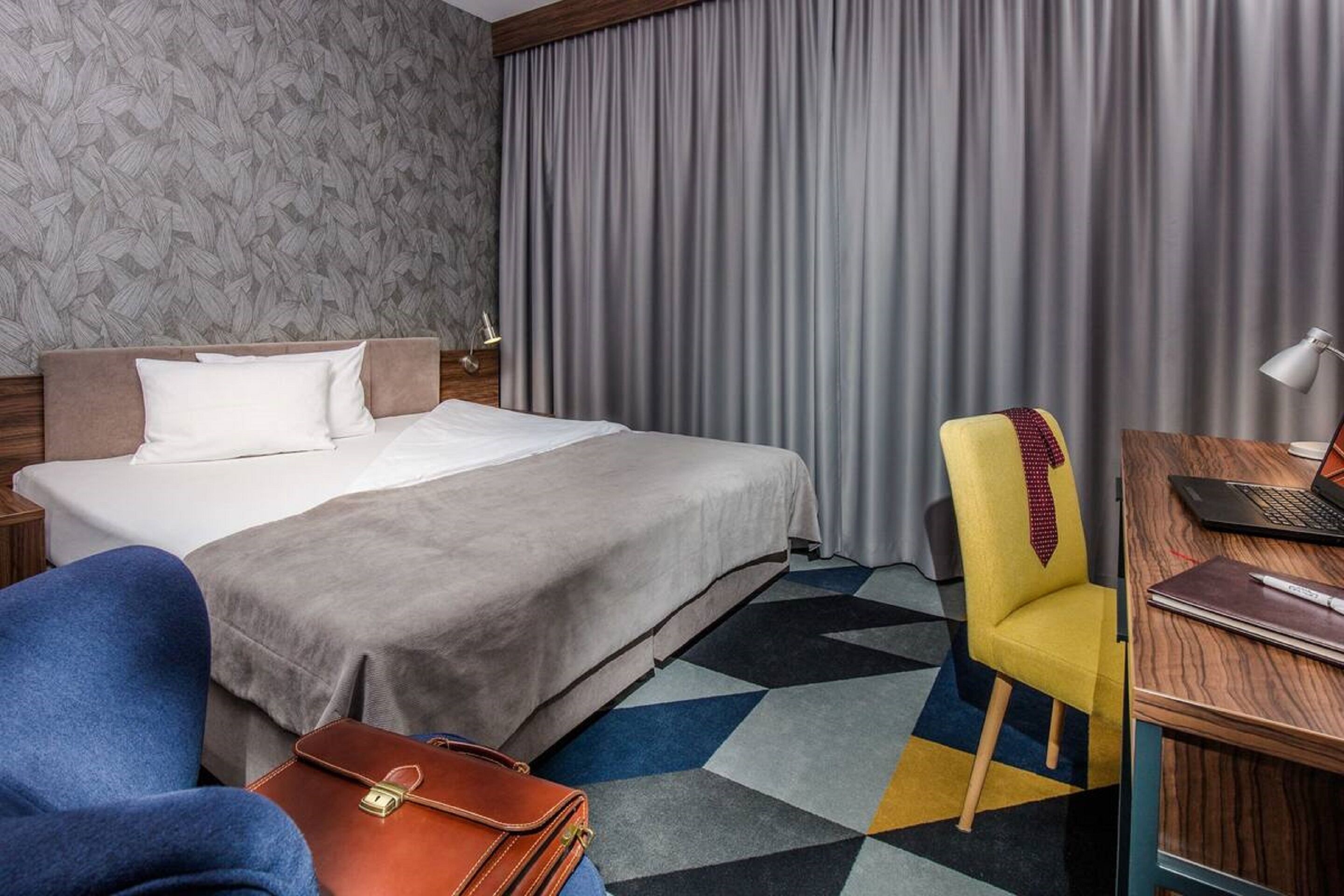 A hotel room with a bed, a desk with a laptop, a chair, and a suitcase.