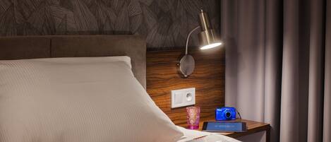 A neatly made bed with a pillow, a bedside table with a lamp, a book, a camera, and a cup.