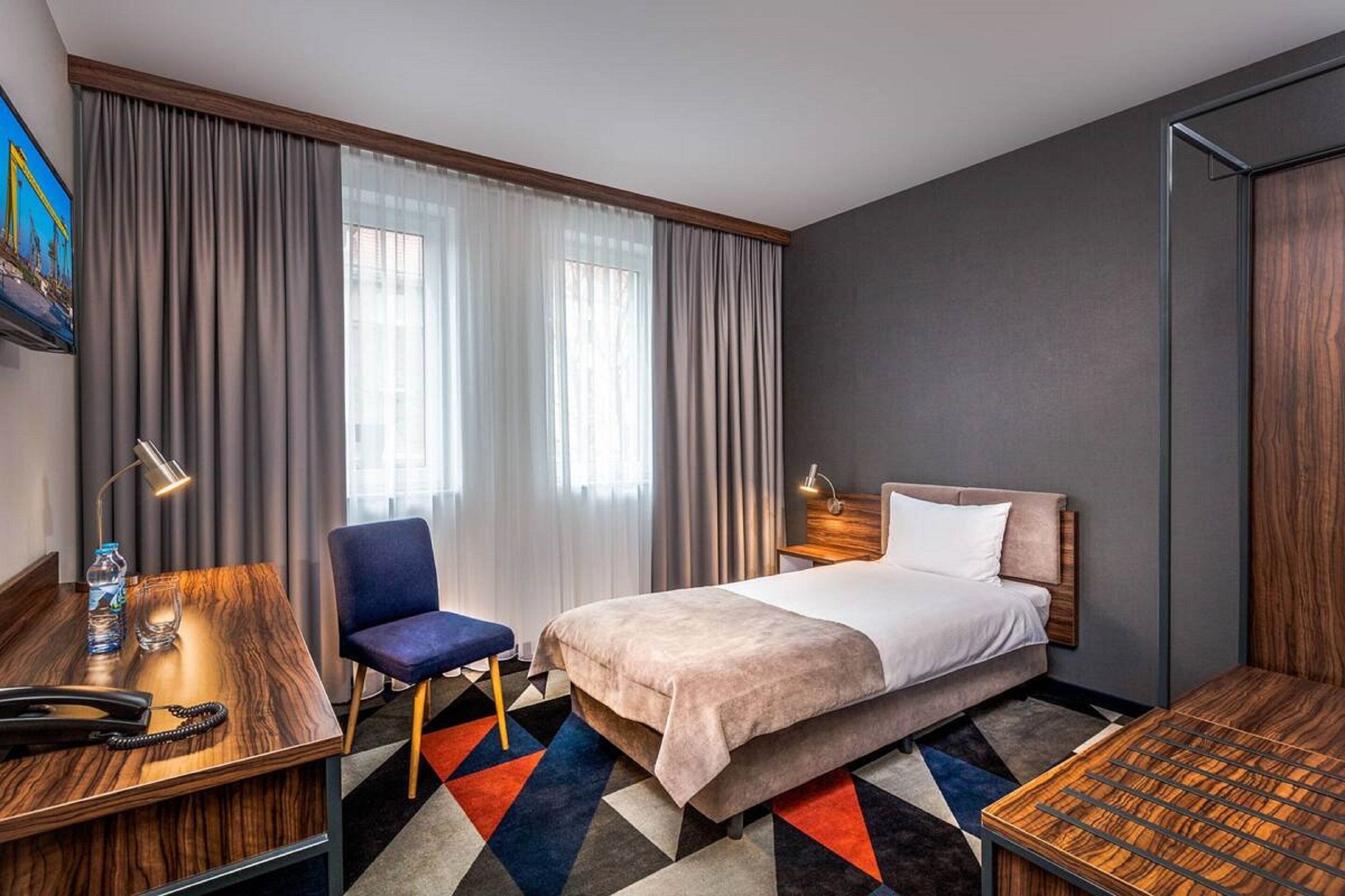 Habitación de hotel con cama, escritorio, silla y ventana grande con cortinas.