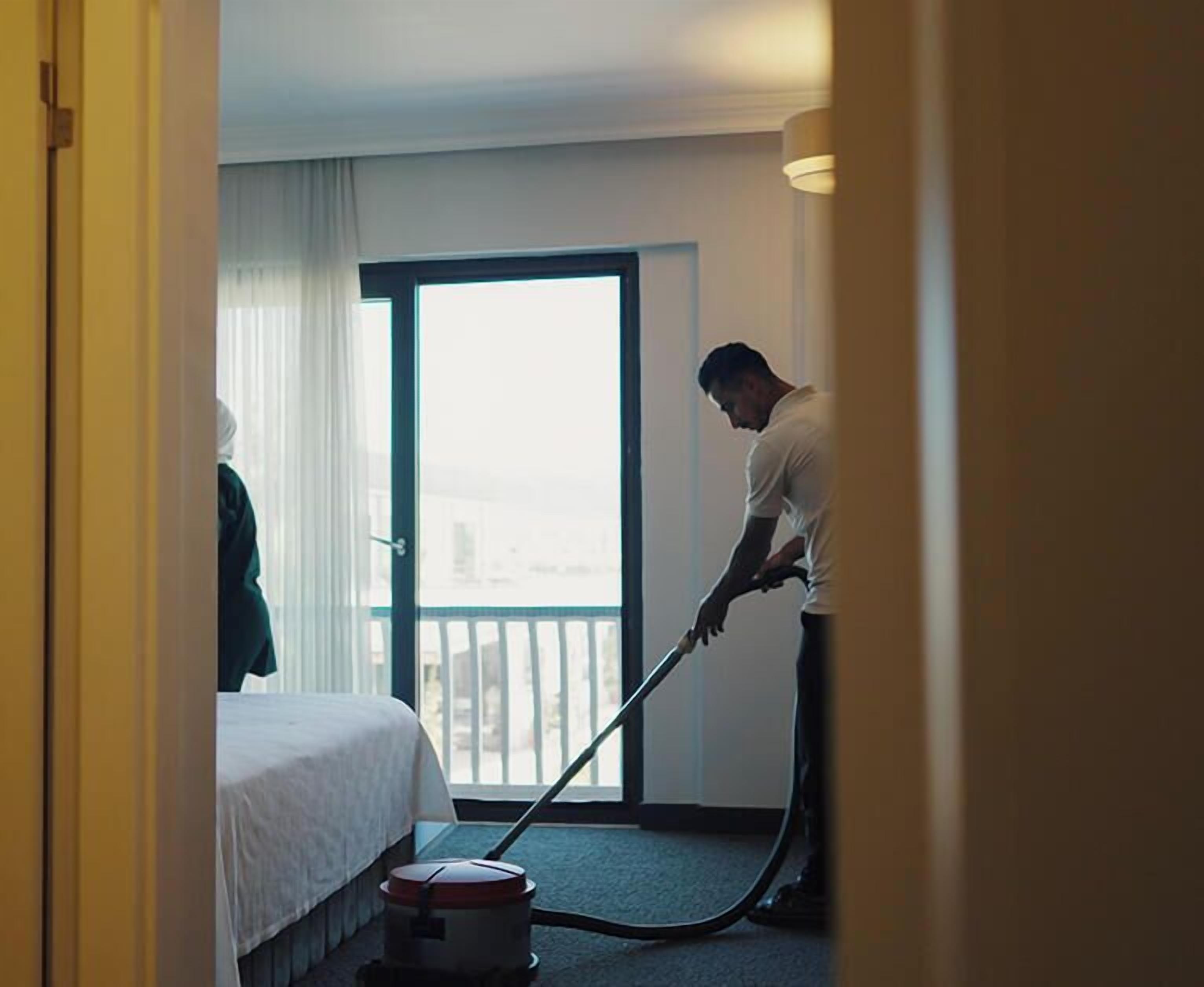 A hotel room with a bed, a window with curtains, and a person vacuuming.