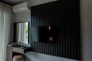 A modern hotel room with a flat-screen TV mounted on a dark wall, a small desk with a remote control, and a window with curtains.