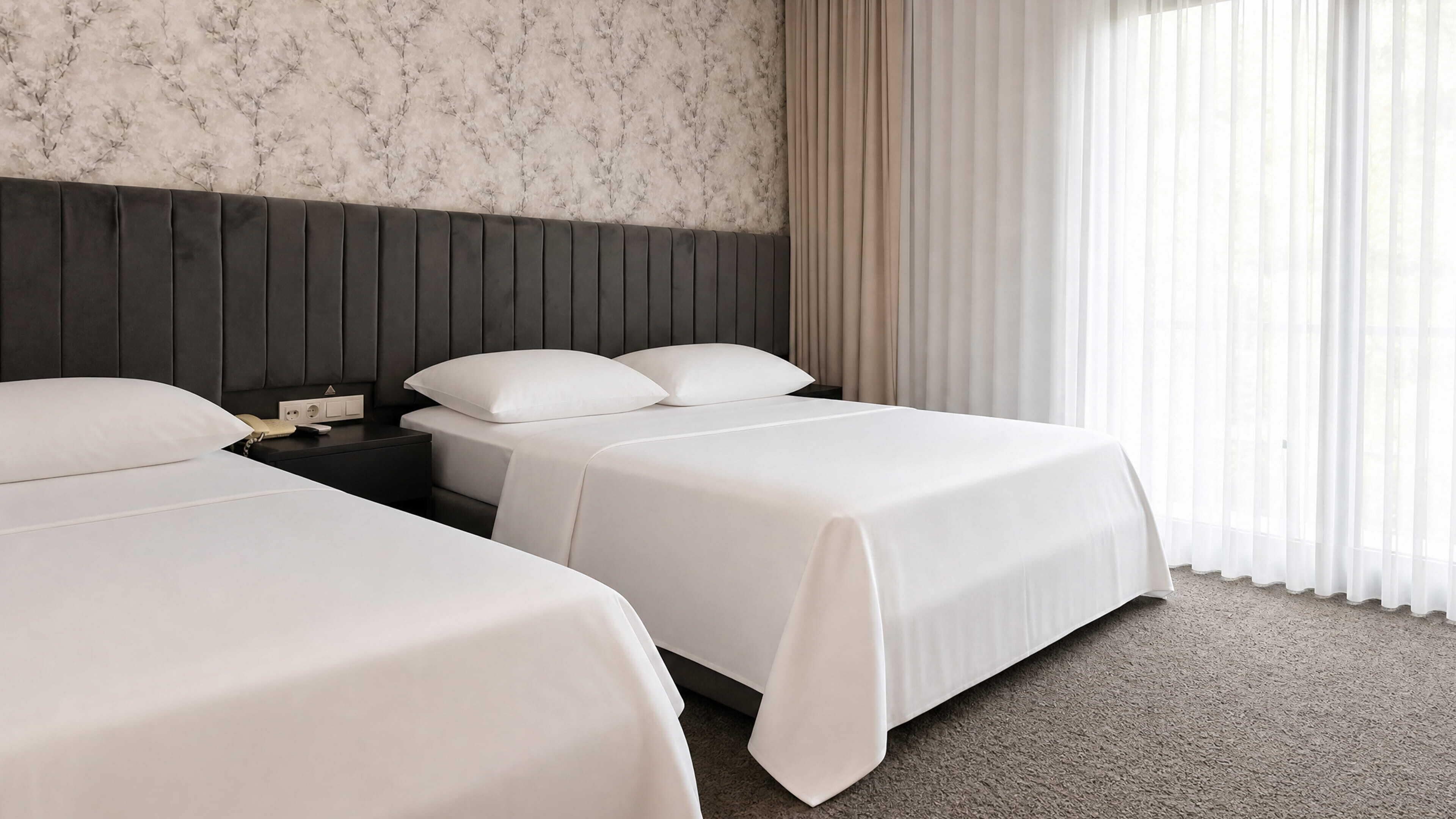 A hotel room with two beds, a dark headboard, a nightstand with a phone, and a window with sheer curtains.