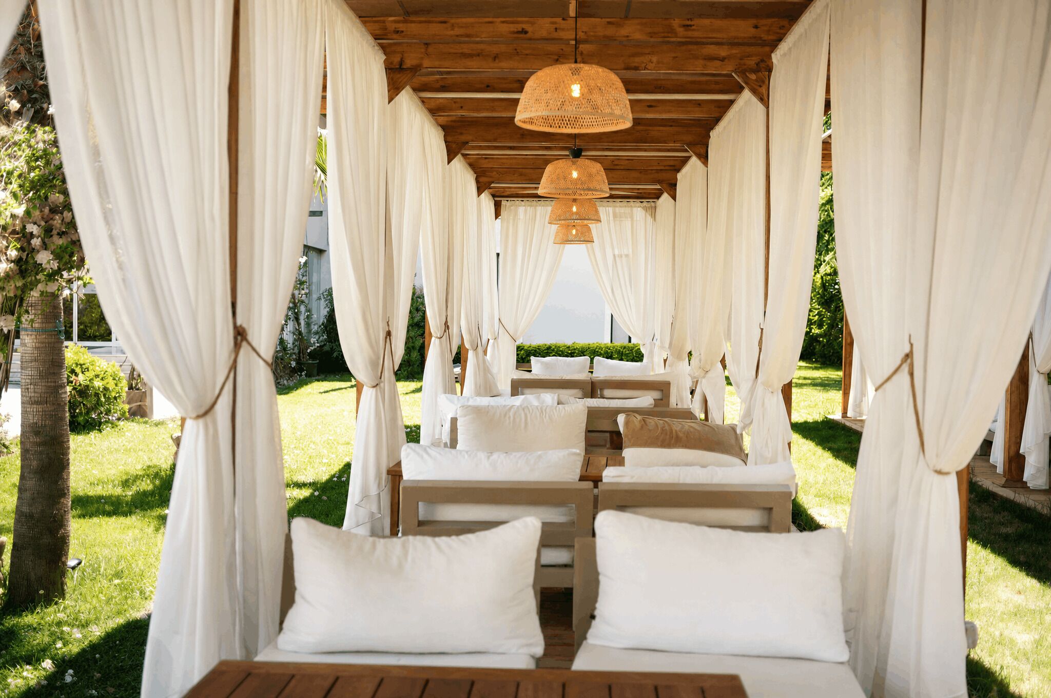 Un área de estar al aire libre con techo, con bancos de madera y cojines blancos, rodeada de cortinas blancas y un techo de madera.