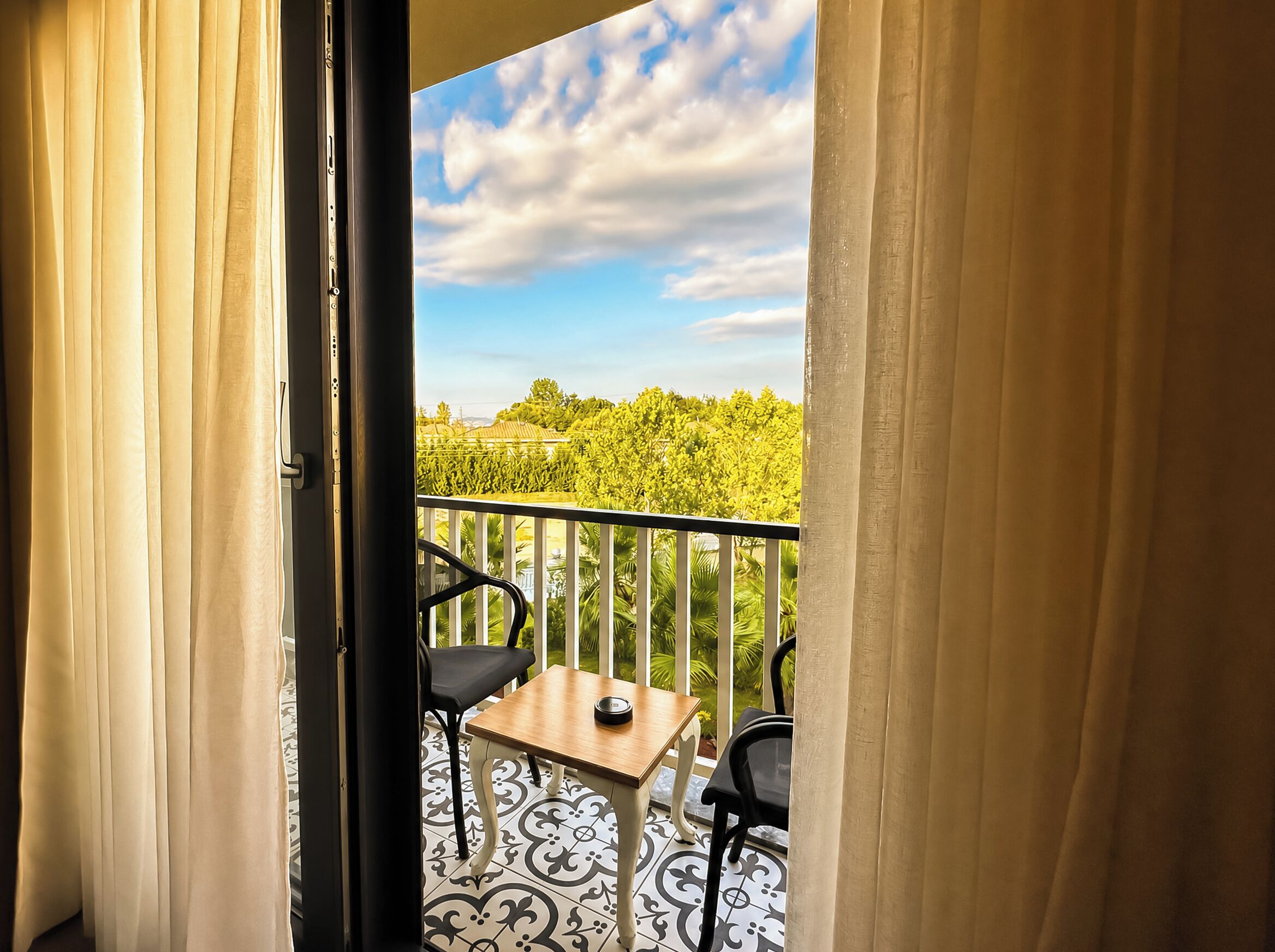 A balcony with a table and chairs, a view of greenery, and a partly cloudy sky.