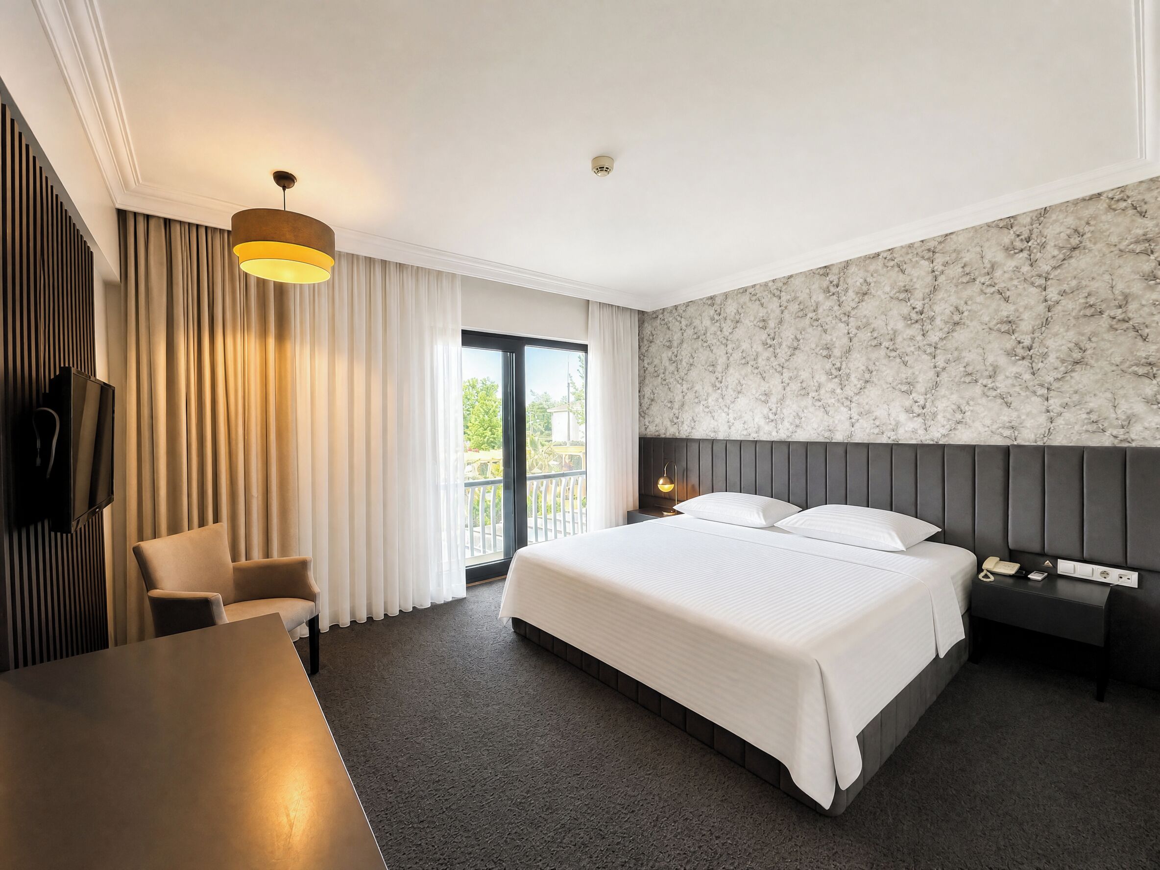 A hotel room with a bed, a desk, a chair, a TV, a window with curtains, and a marble-patterned wall.