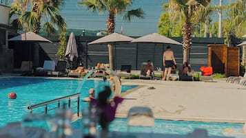 Zona de piscina con sillas de descanso, sombrillas y palmeras.