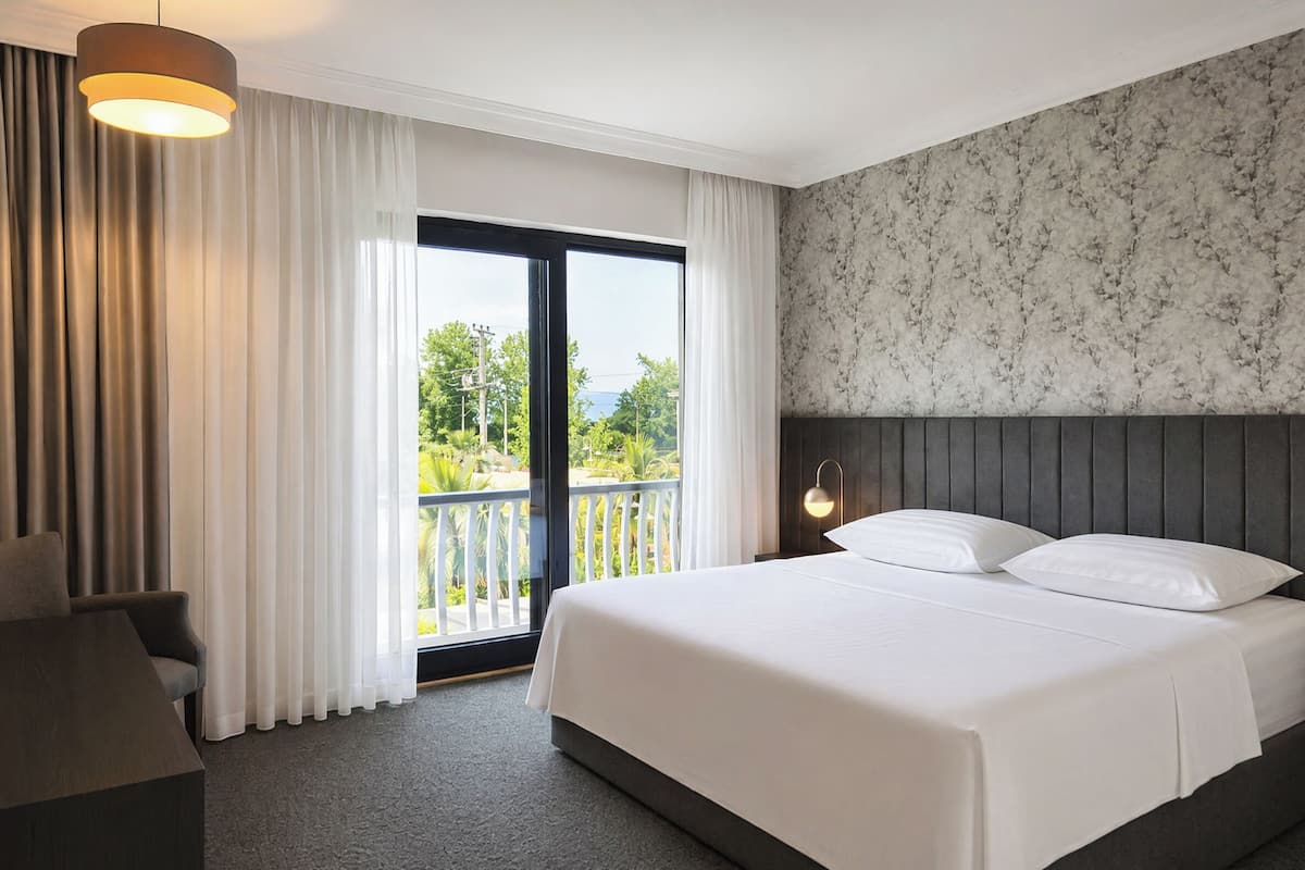Habitación de hotel con cama, una silla, balcón con cortinas y vista a la vegetación.