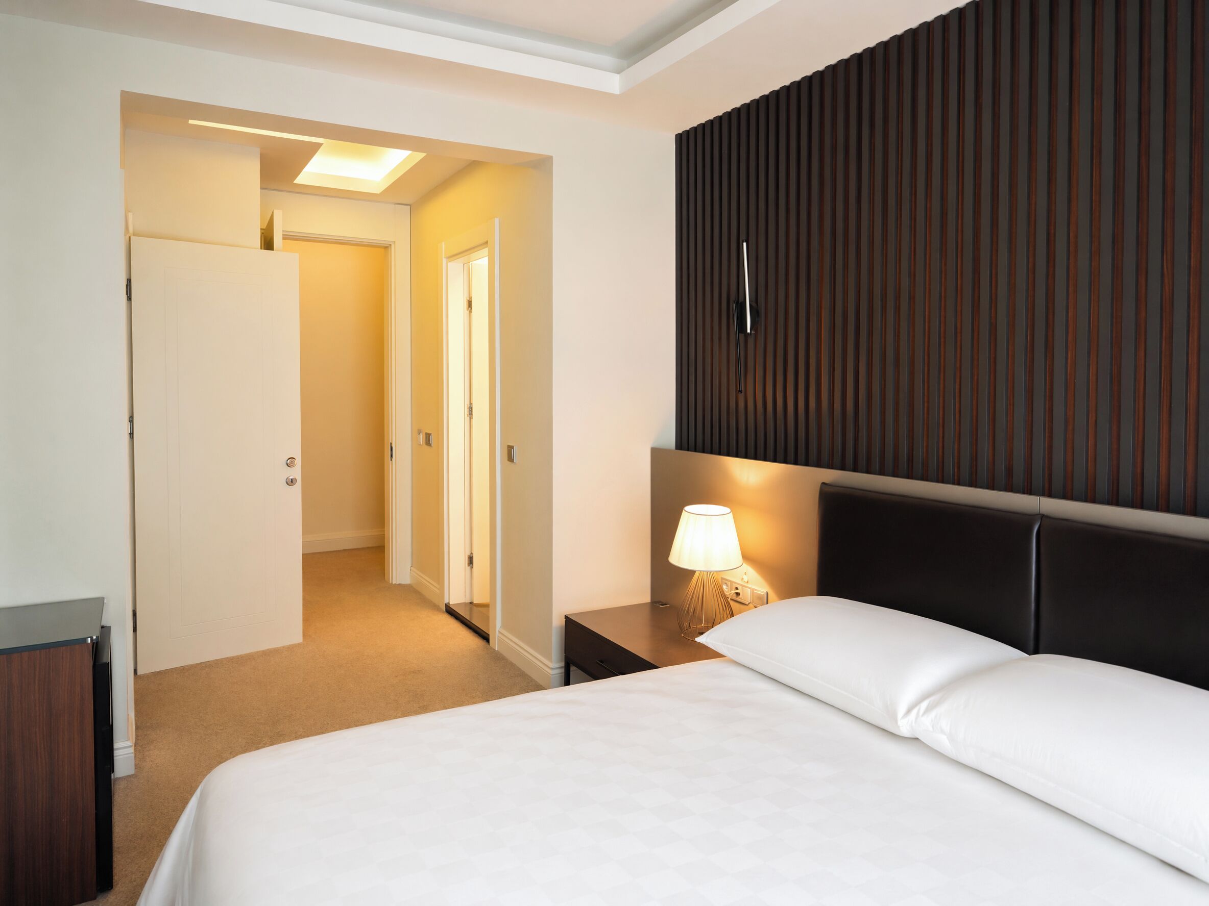 Une chambre d’hôtel avec un lit, une table de chevet avec une lampe, une tête de lit en bois et une porte donnant sur une autre pièce.