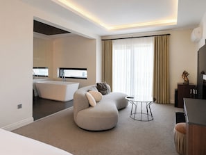 A modern hotel room with a curved sofa, a small round table, a bathtub, and a large window with curtains.