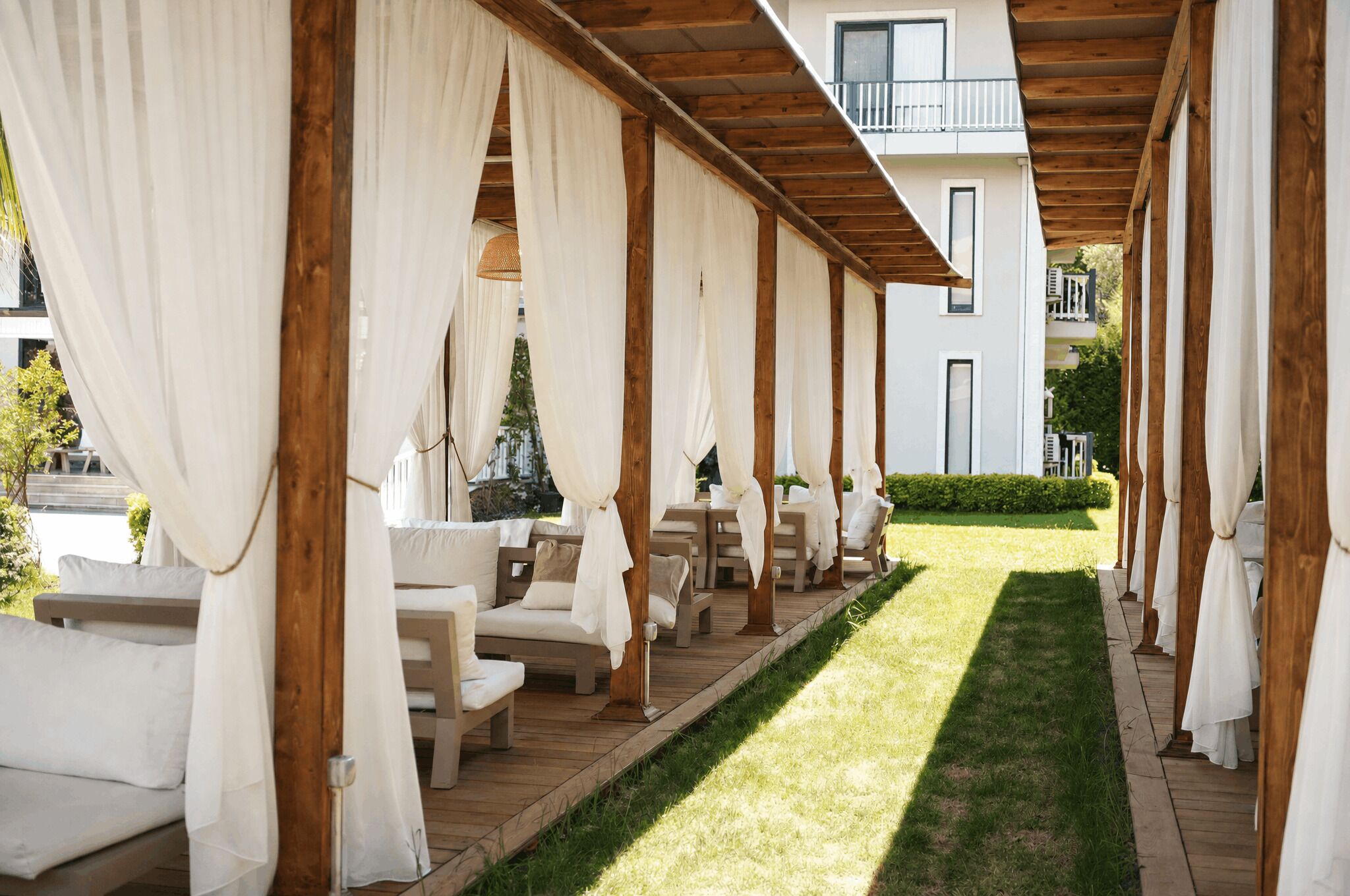 Un espacio de estar al aire libre cubierto con pilares de madera y cortinas blancas, un césped y un edificio al fondo.