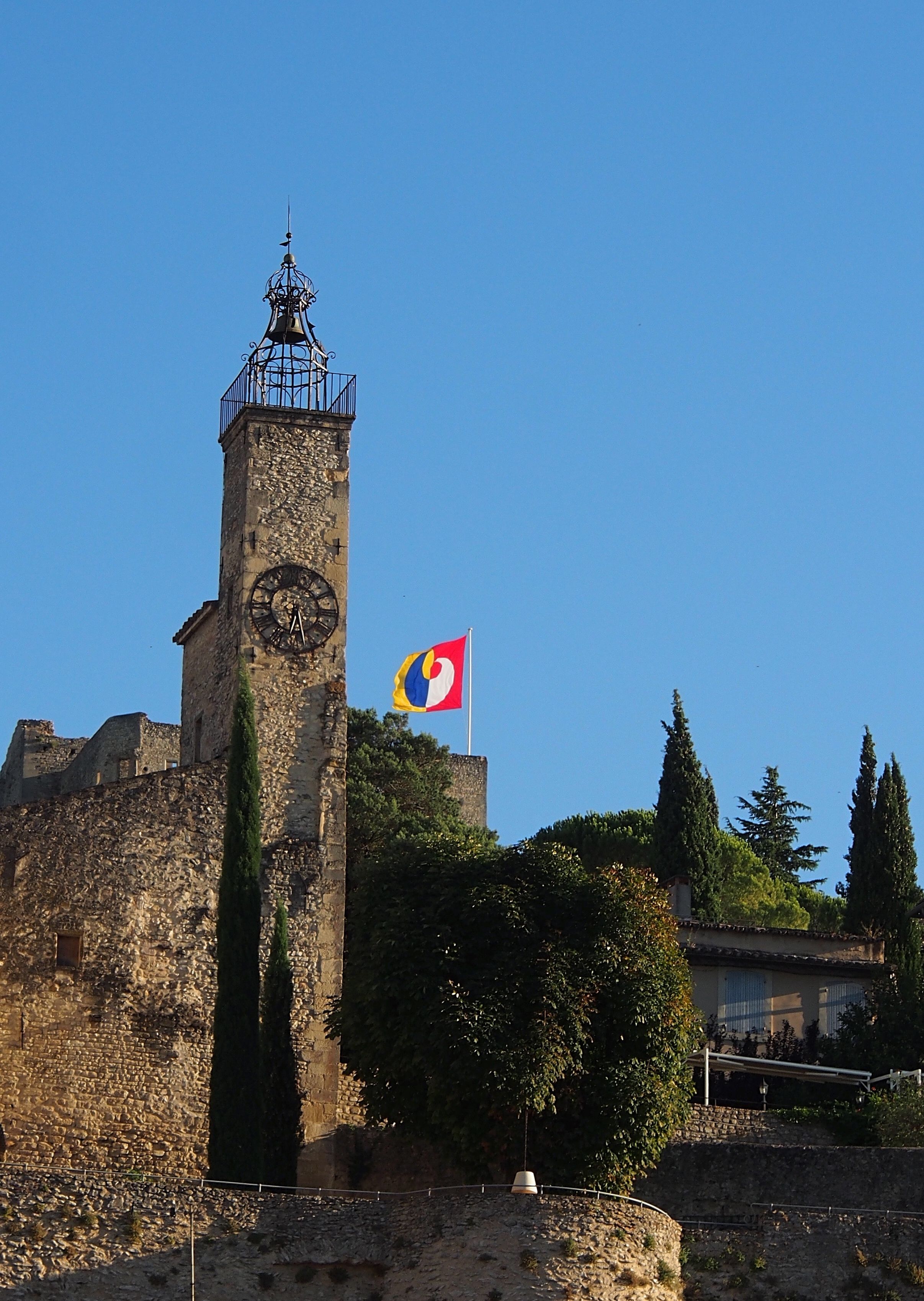A historic stone tower with a clock, a flag, and surrounding greenery.