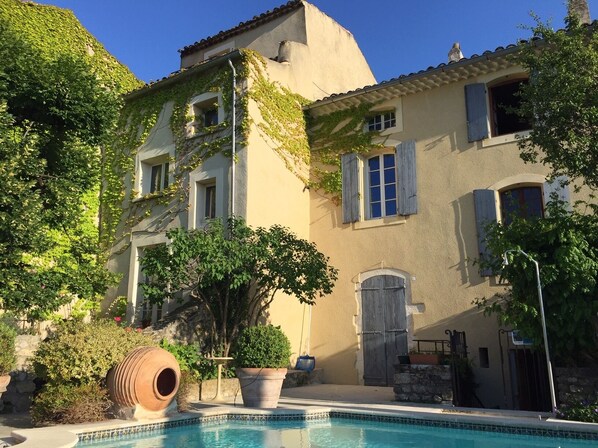 A poolside view of a Mediterranean-style building with a pool, potted plants, and a rustic barrel.