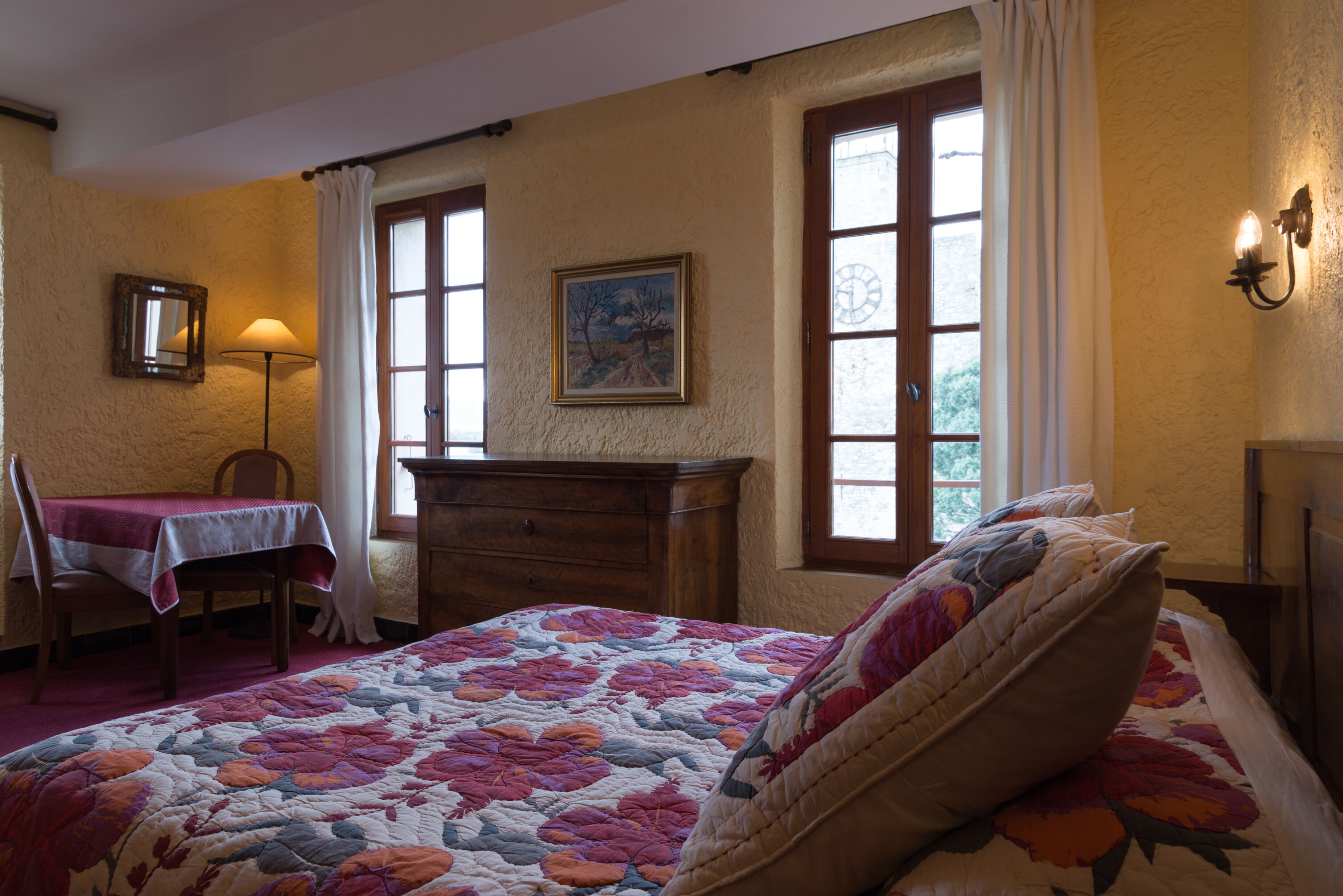 Un dormitorio con una colcha de patchwork floral, un aparador de madera, una silla con una mesa y un cuadro en la pared.