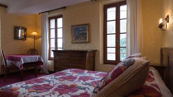 A bedroom with a floral quilted bedspread, a wooden dresser, a chair with a table, and a painting on the wall.