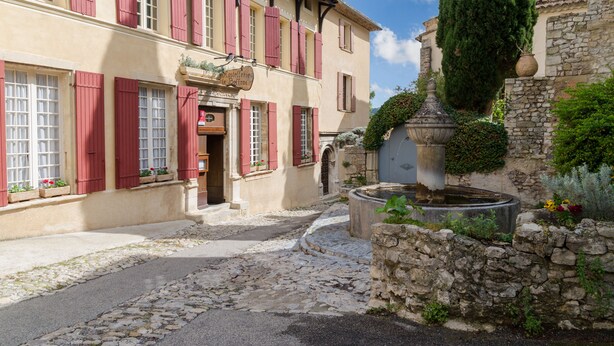 A traditional building with red shutters, a stone fountain, and a cobblestone street. - Hostellerie Le Beffroi (Vaison-la-Romaine)