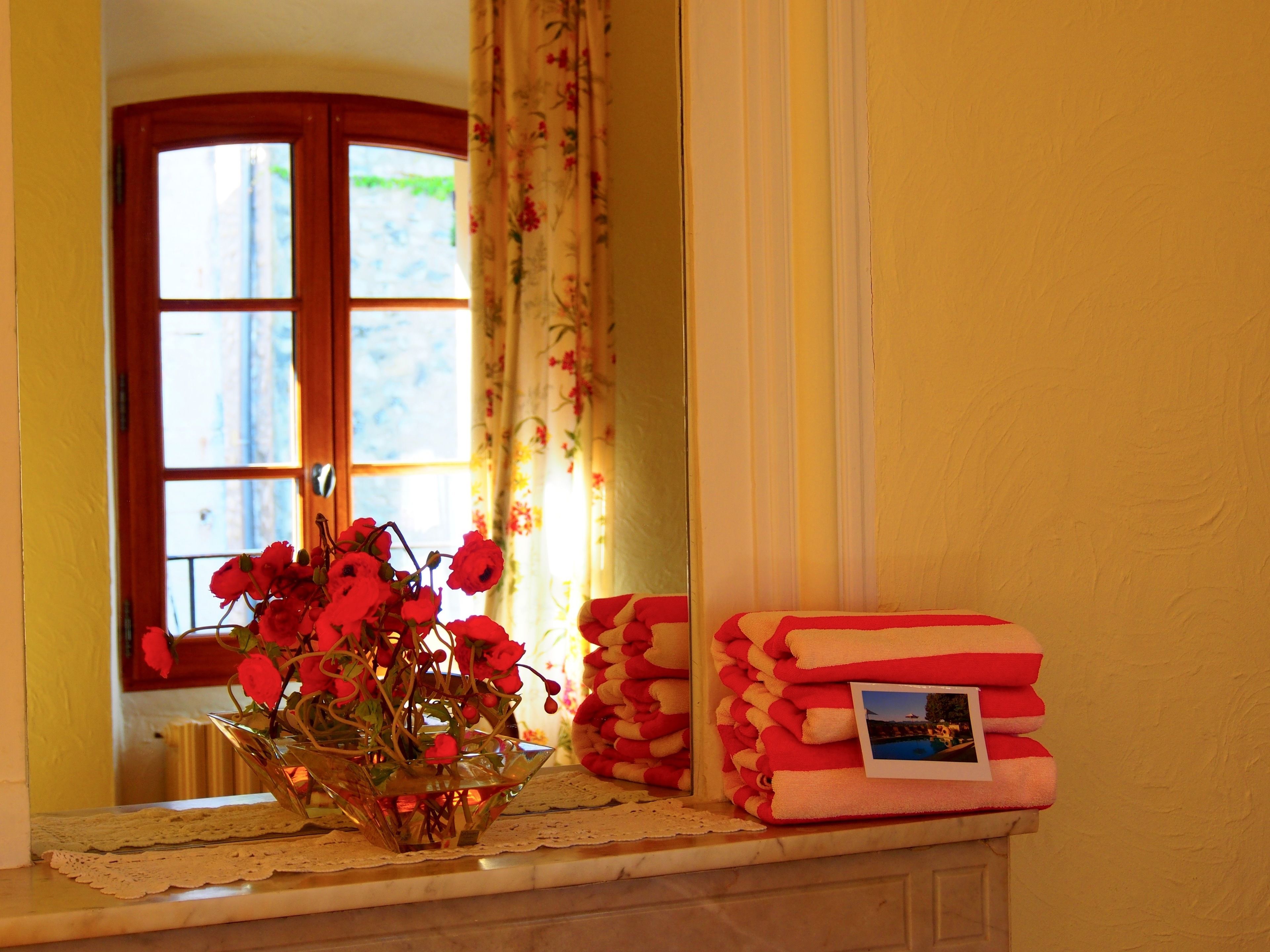 A window with floral curtains, a vase with red flowers, and stacked towels with a photo on them.