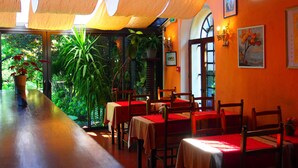 An indoor dining area with tables and chairs, a large window with a view of greenery, and artwork on the walls. - Hostellerie Le Beffroi (Vaison-la-Romaine)