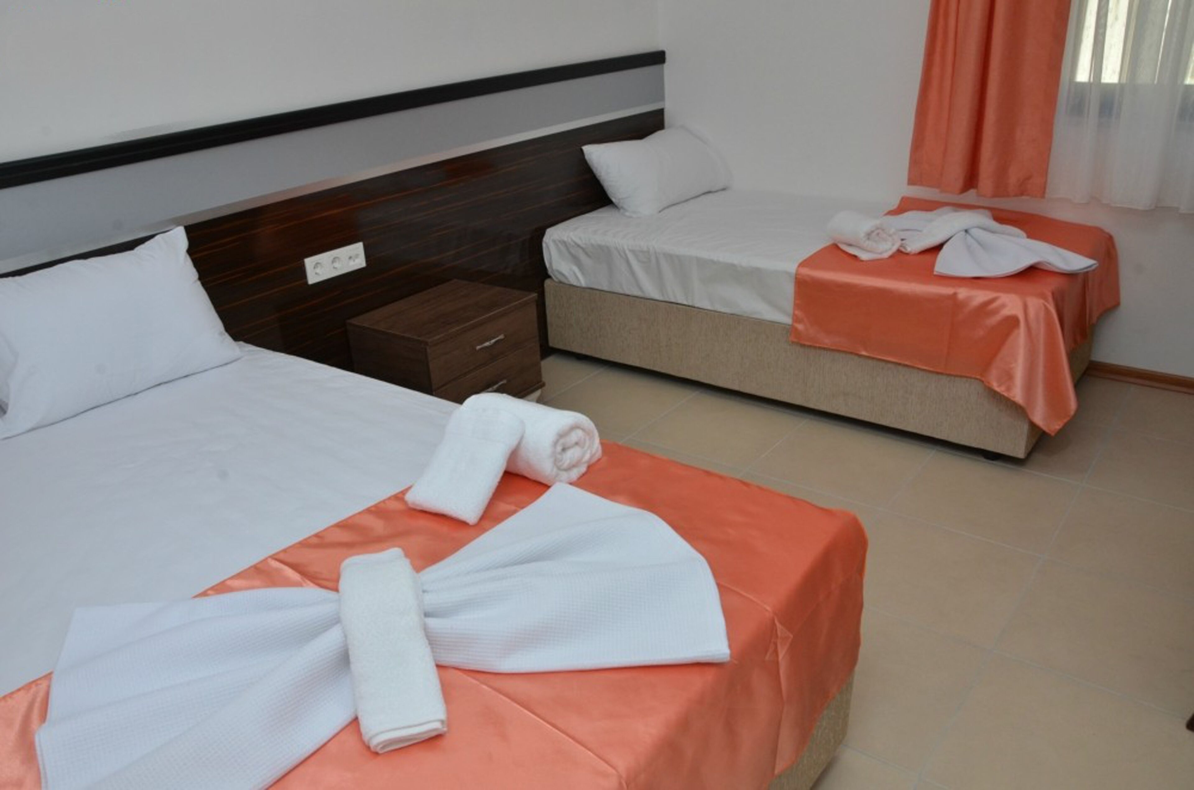 Habitación de hotel con dos camas, cada una con cubrecama blanco y manta naranja. Hay toallas blancas sobre las camas y una mesita de noche de madera con un cajón.