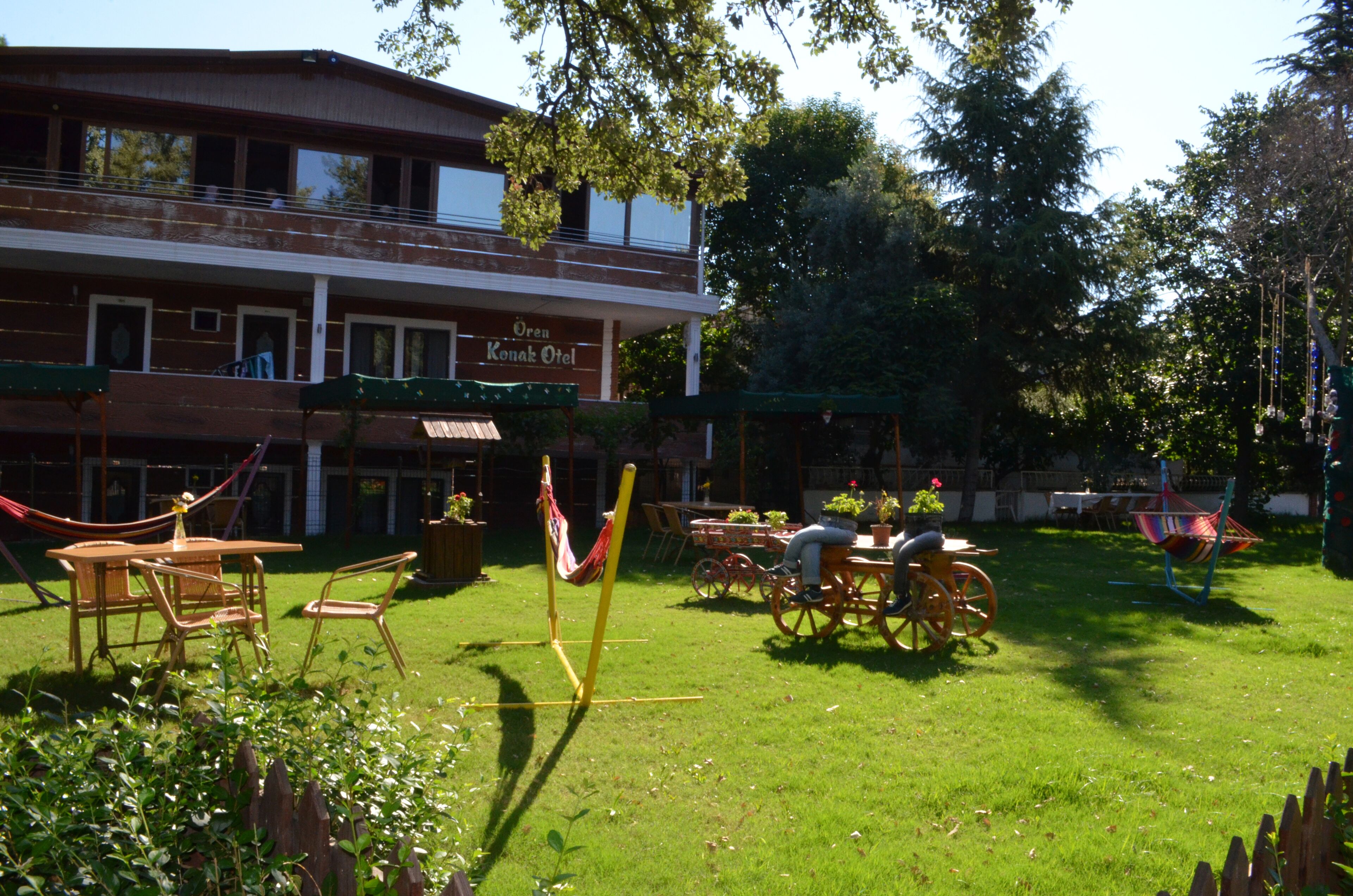 A building with a sign reading "Oren Tak Hotel" and a grassy outdoor area with tables, chairs, and hammocks.