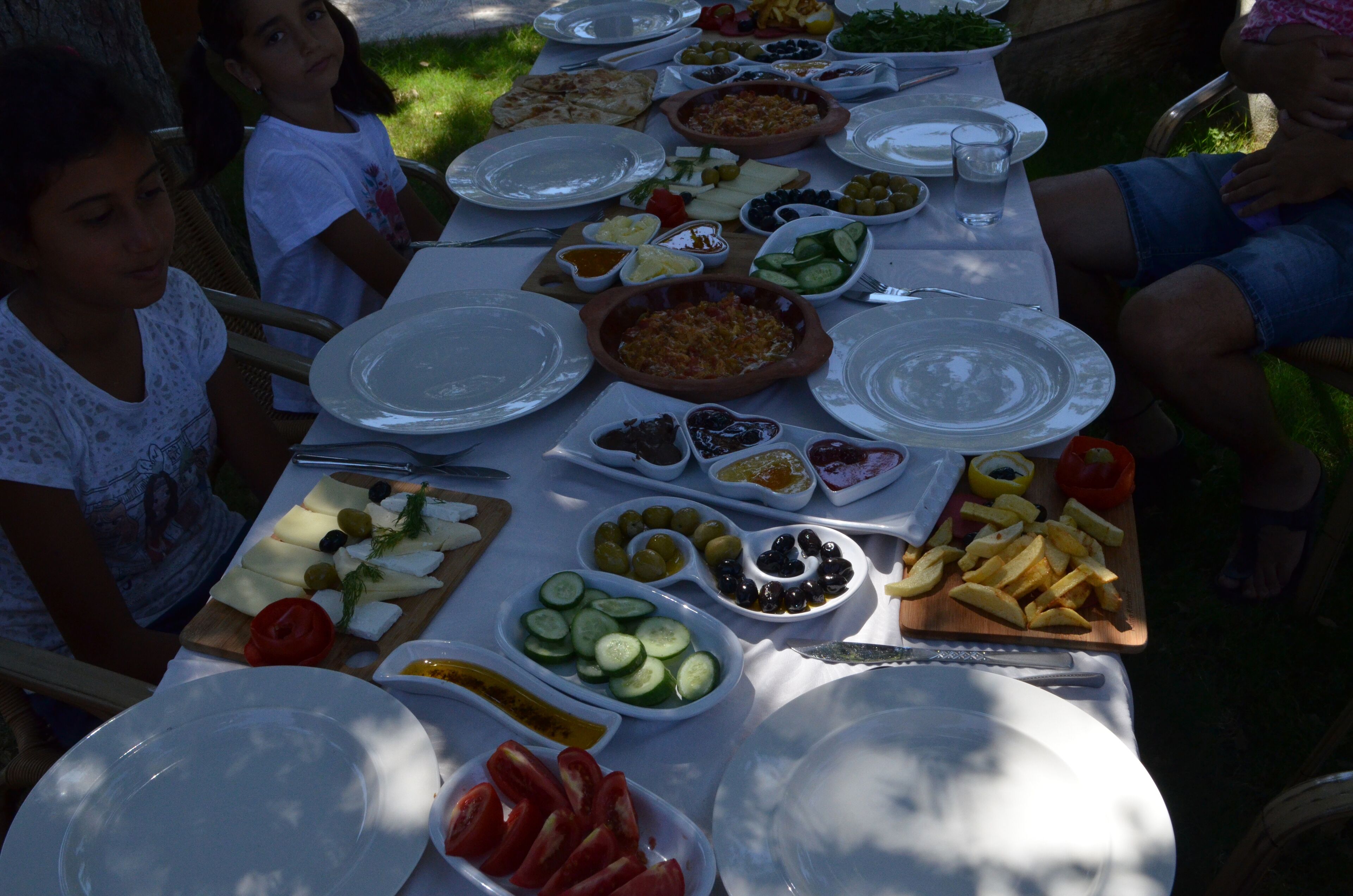 Mesa posta ao ar livre com diversos pratos, incluindo azeitonas, tomates, pepinos e pão.