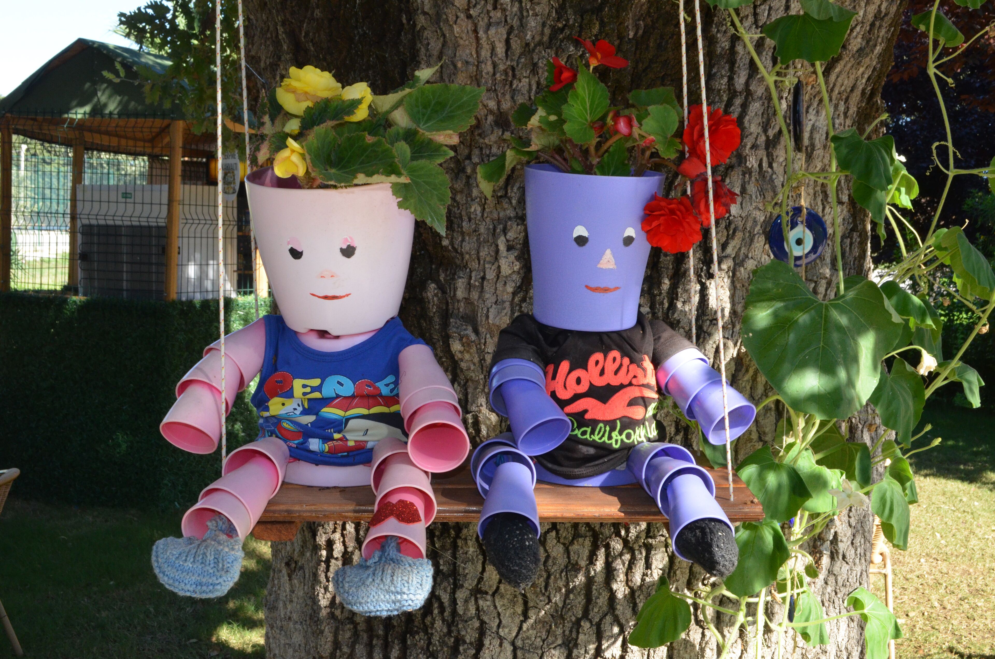 Two large potted plants with face-like decorations on a wooden bench against a tree.