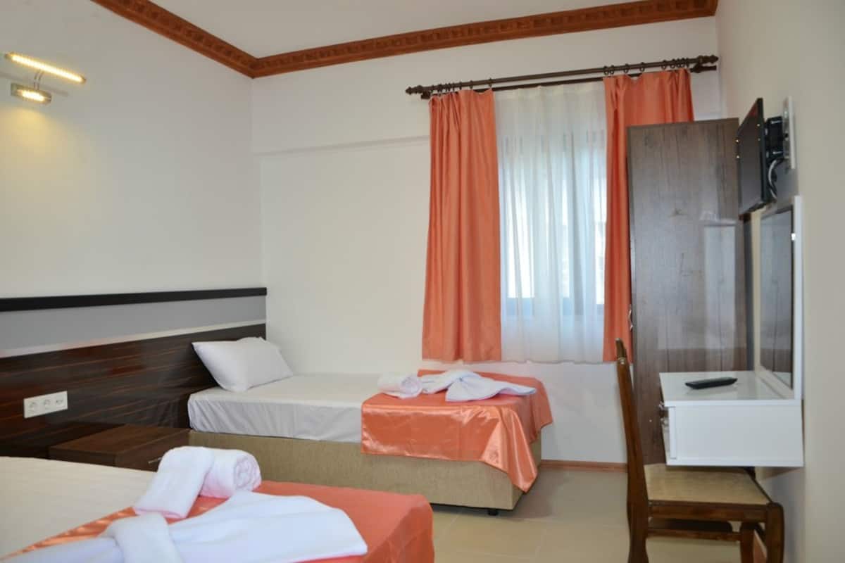 A hotel room with two single beds, a wooden headboard, a small table with a remote control, a mirror, and a window with orange curtains.