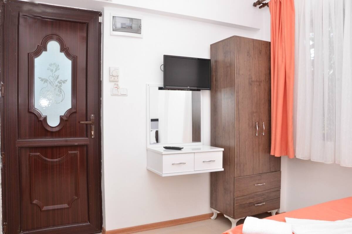A bedroom with a wooden wardrobe, a white bedside table, a TV mounted above the table, and a bed with an orange bedspread.
