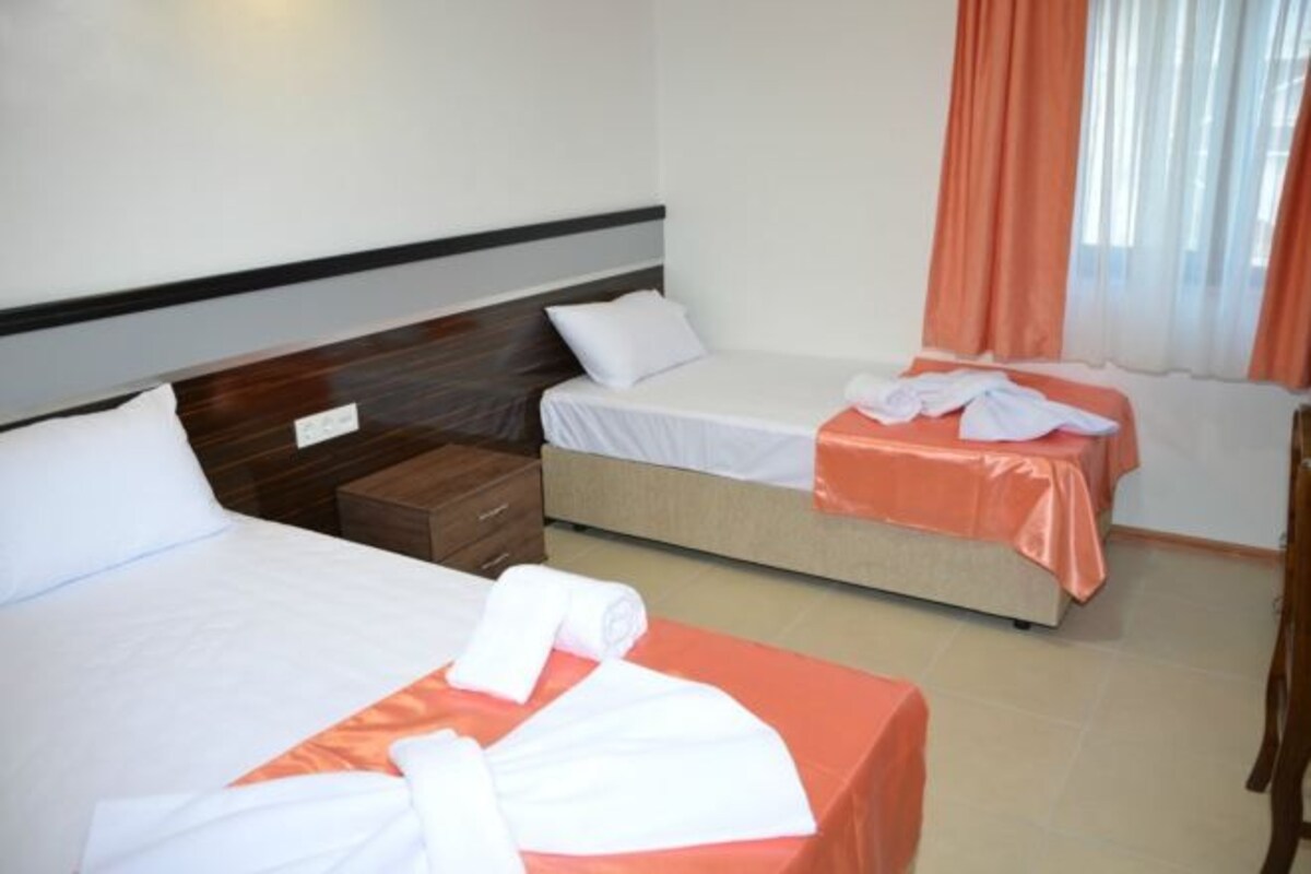 A hotel room with two beds, each with a white bedspread and orange curtains. There is a wooden nightstand between the beds, and a small table with a chair to the right.