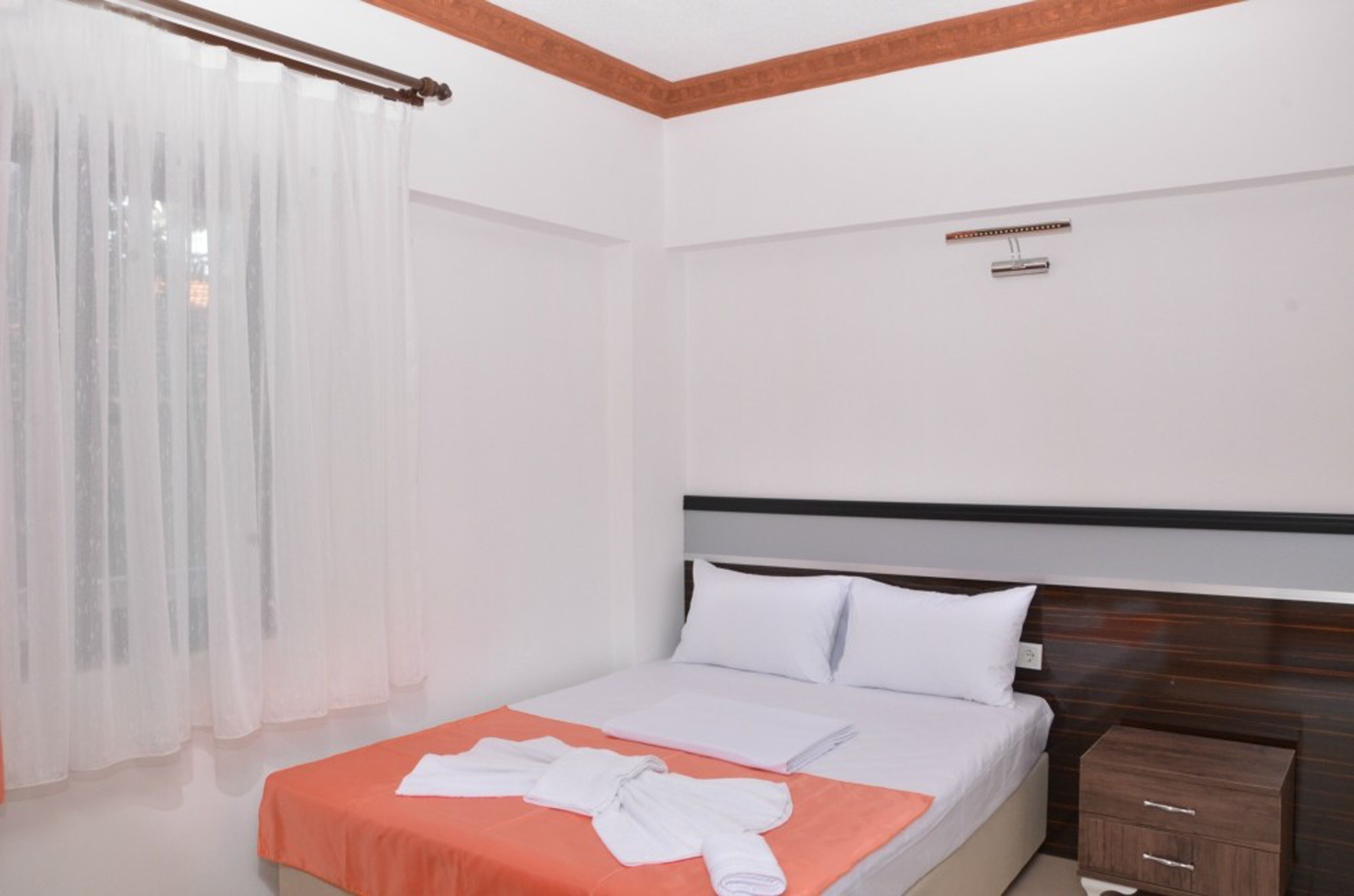 Una habitación de hotel con cama, mesita de noche, cabecera de madera y ventana con cortinas translúcidas.