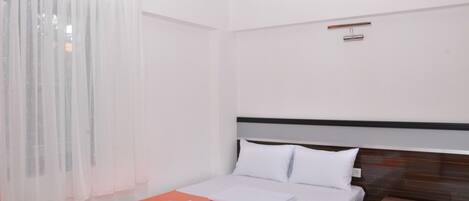 Una habitación de hotel con cama, mesita de noche, cabecera de madera y ventana con cortinas translúcidas.