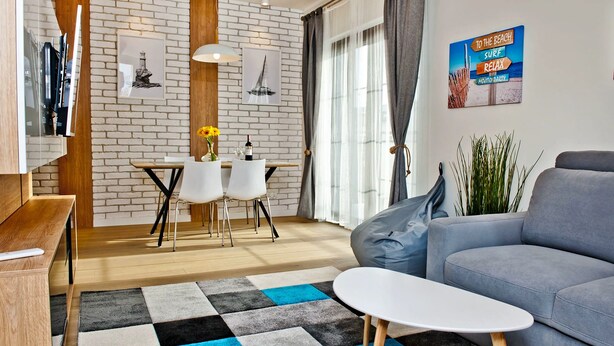 A modern living room with a grey sofa, a coffee table, and a dining area with a wooden table and chairs. There is a brick wall with a wooden accent, a hanging light fixture, and a poster on the wall. - Willa Haven BlueApart Jastarnia (Jastarnia)
