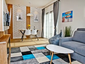 Ein modernes Wohnzimmer mit grauem Sofa, Couchtisch und Essbereich mit Holztisch und Stühlen. Eine Ziegelwand mit Holzelement, eine Pendelleuchte und ein Poster sind ebenfalls zu sehen.