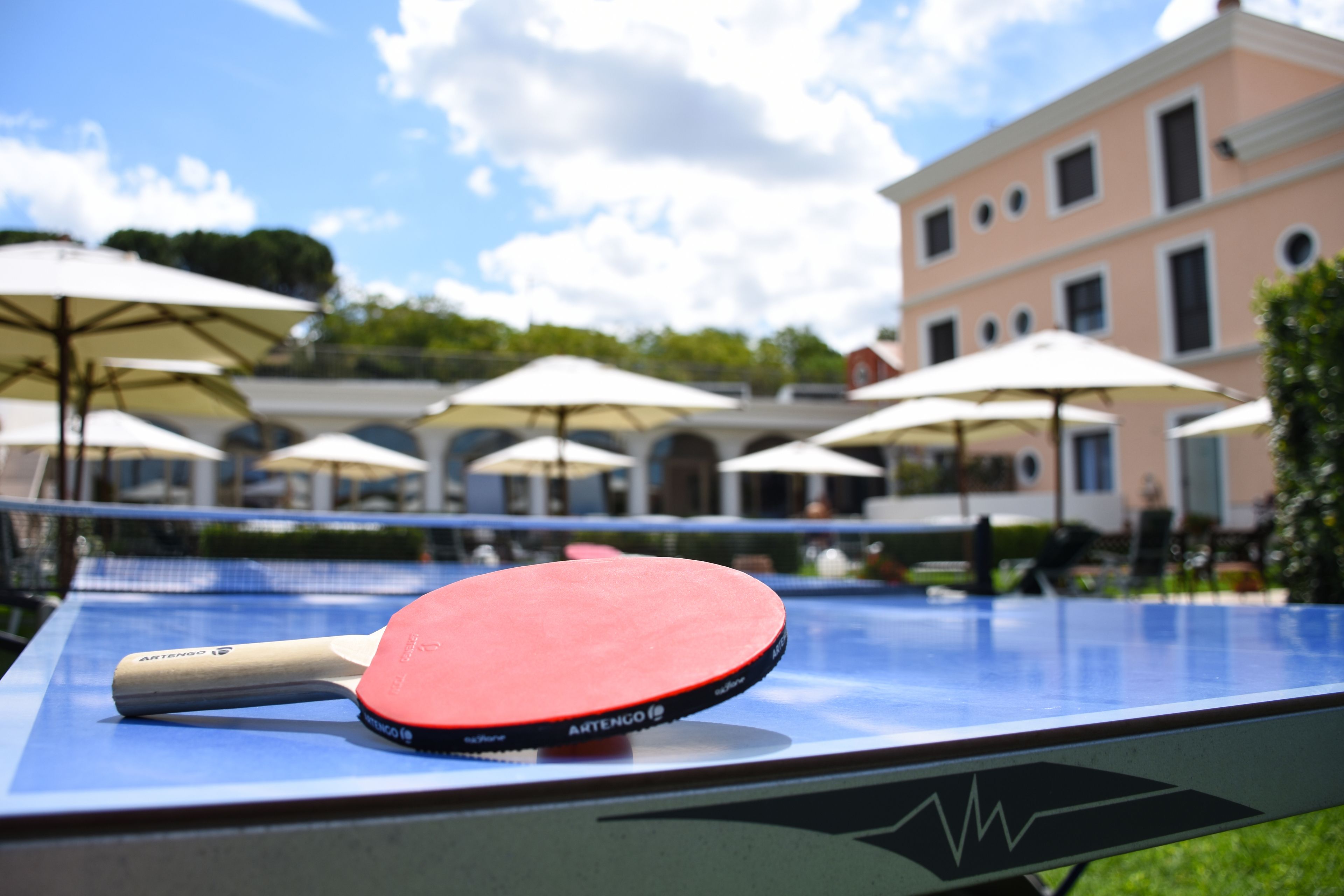 A table tennis table with a paddle, surrounded by umbrellas, with a building in the background.
