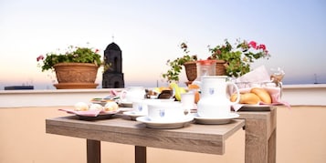 Uma mesa de madeira com pratos, copos e um jarro em um terraço com plantas em vasos e uma igreja ao fundo.