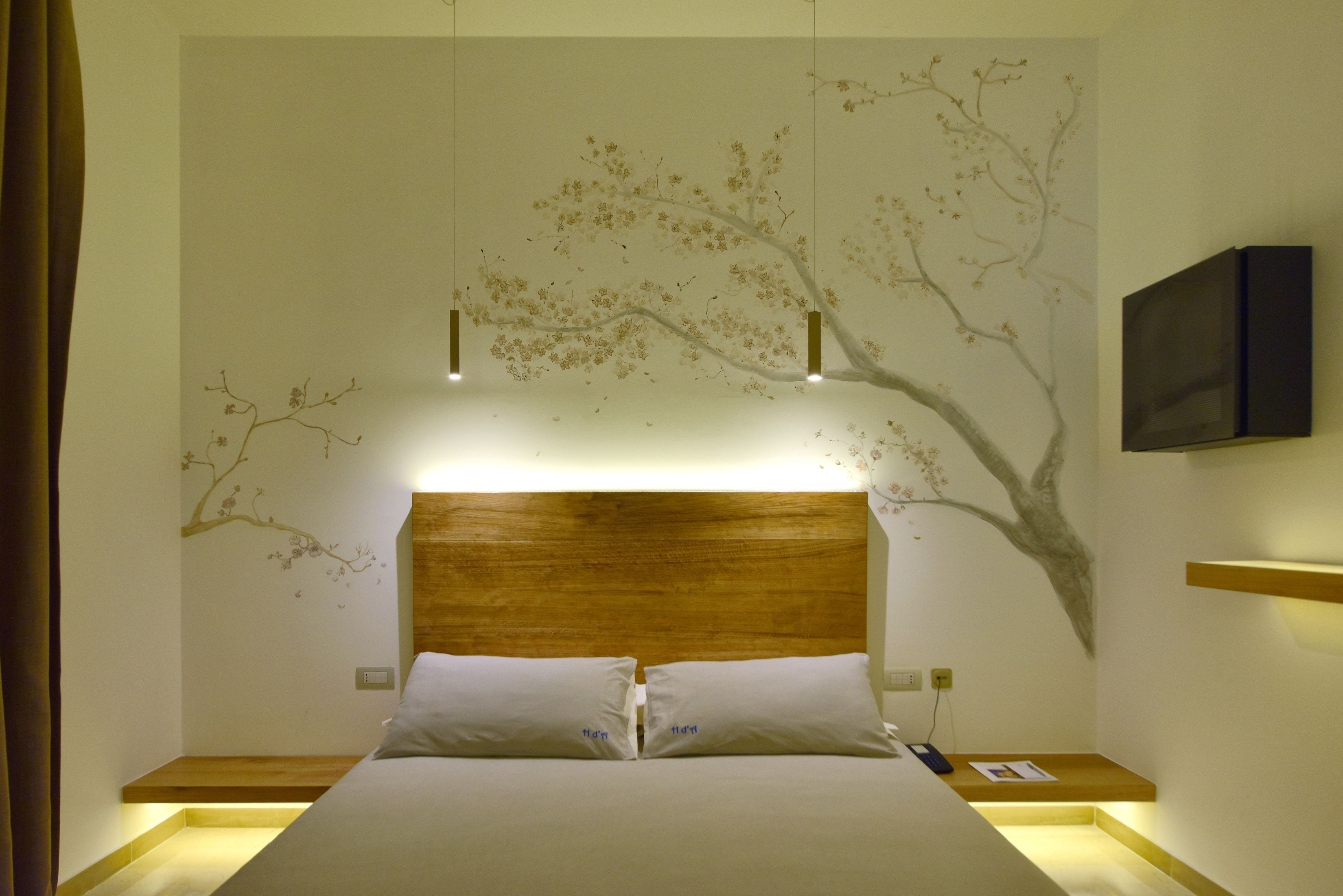 A neatly made bed with a wooden headboard, a wall-mounted TV, and a wall mural of a tree.
