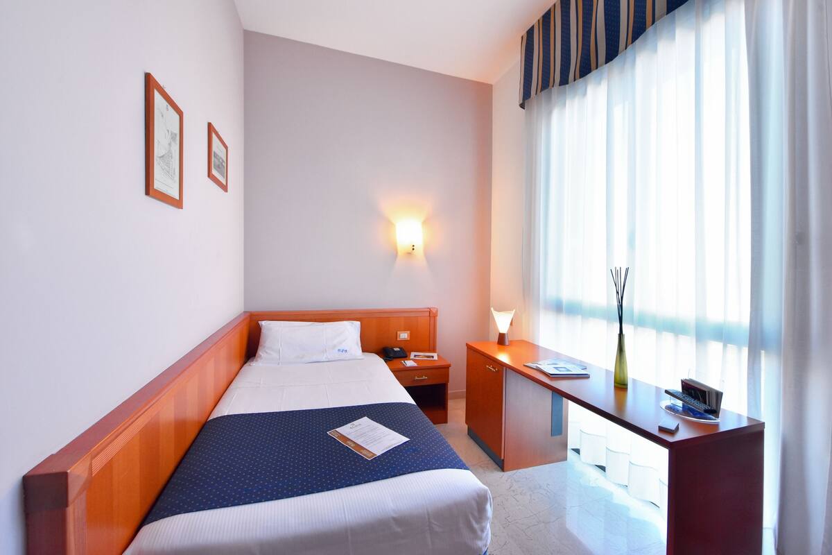 A hotel room with a bed, a desk, a nightstand, a lamp, and a window with curtains.