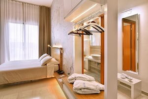 Quarto de hotel moderno com uma cama, banheiro com pia e espelho, e um cabideiro com roupas.
