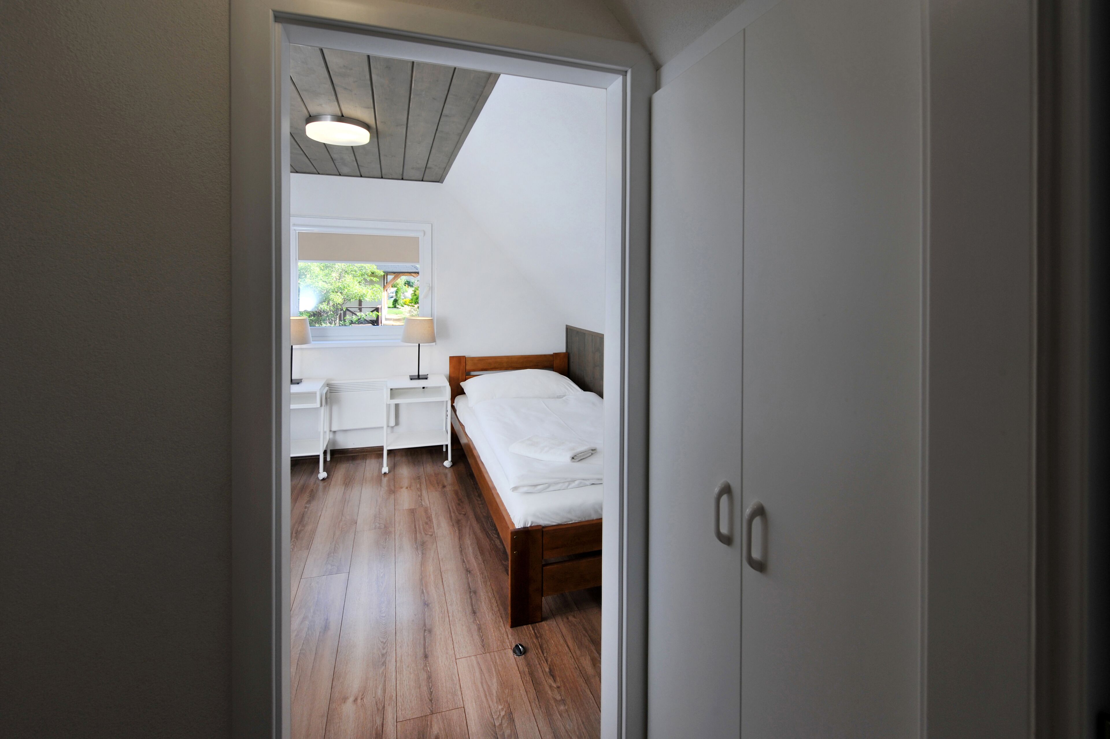 A bedroom with a wooden bed, a bedside table, a nightstand, and a window with a view of greenery.