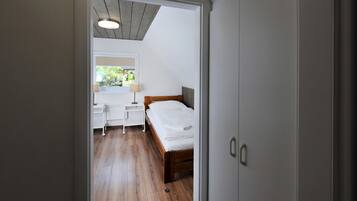 A bedroom with a wooden bed, a bedside table, a nightstand, and a window with a view of greenery.
