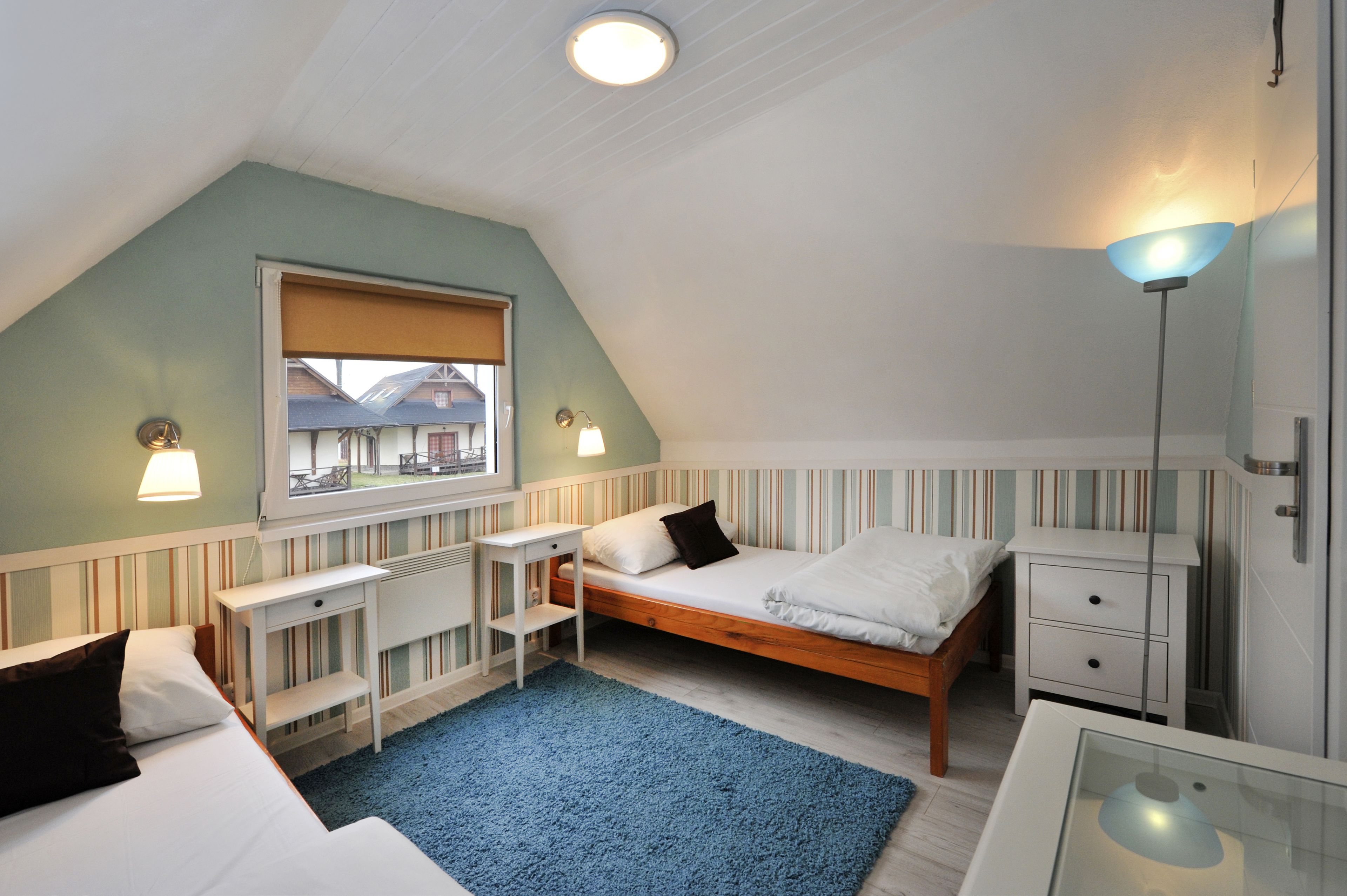 Un dormitorio con una cama individual, un escritorio, una silla, una mesita de noche y una ventana con vistas a casas.