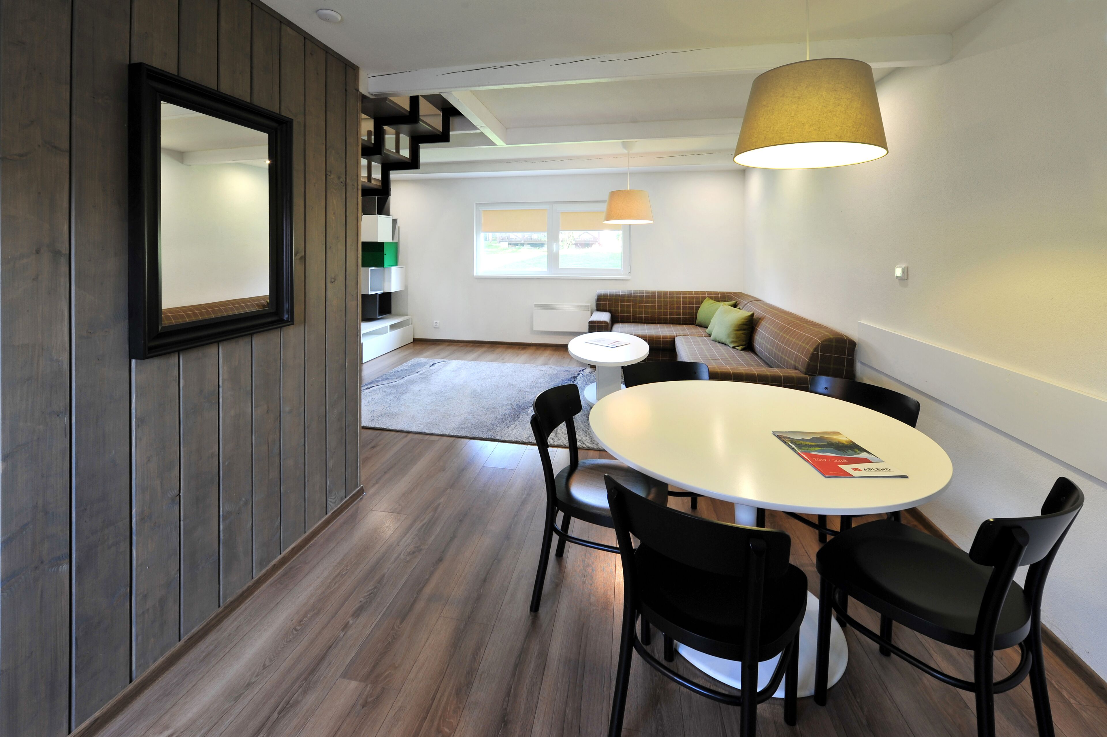 Un salon moderne avec une table ronde, des chaises noires, un canapé avec des coussins, un grand miroir et une lampe suspendue.