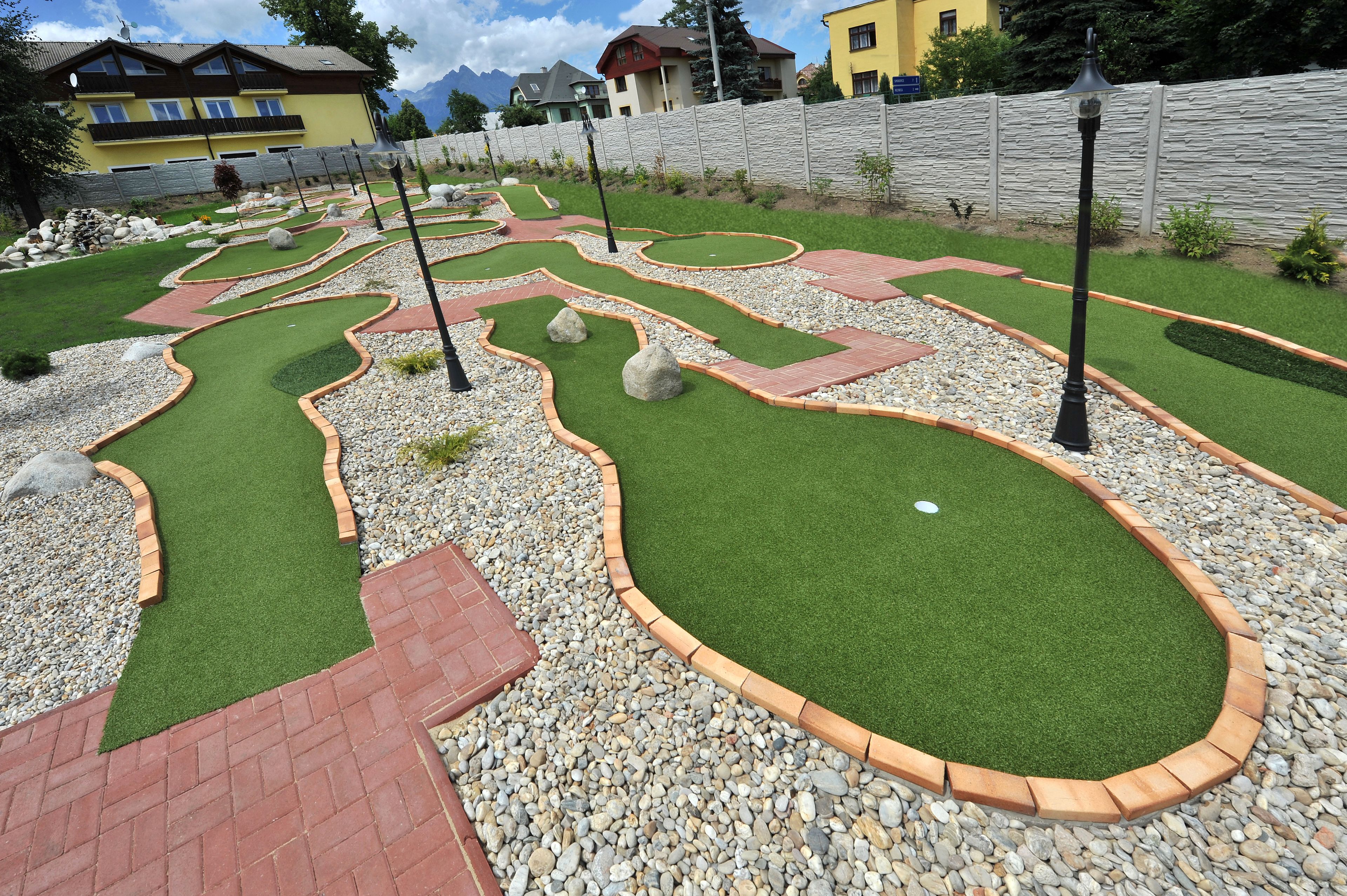 A miniature golf course with green fairways, red brick pathways, and a stone wall in the background.