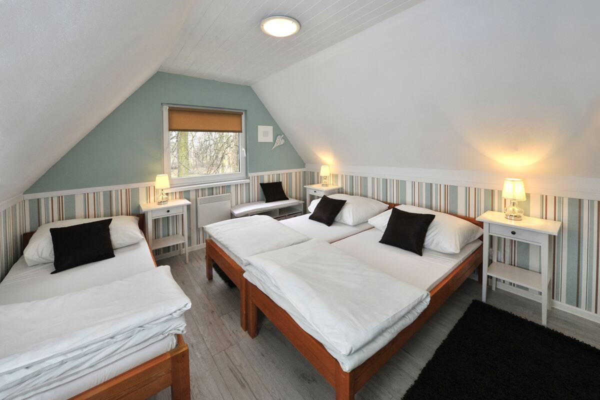 Ein Zimmer mit drei Betten, jeweils mit weißer Bettwäsche und schwarzen Kissen, Holz-Kopfteilen und gestreifter Tapete. Es gibt Nachttische mit Lampen sowie ein Fenster mit Jalousie.