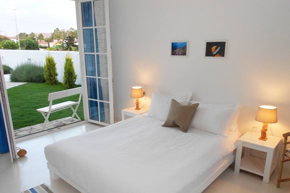 Un dormitorio con una cama grande, dos mesitas de noche con lámparas, una ventana con vistas a zonas verdes y dos pequeños cuadros en la pared.