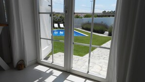 Quarto com uma grande porta de correr de vidro que dá para um quintal com piscina, gramado e cerca branca.