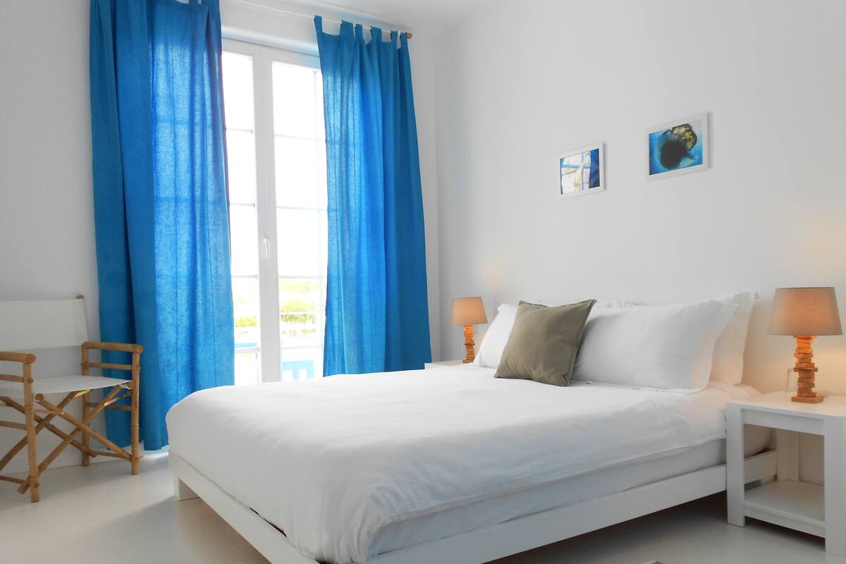 Un dormitorio blanco con una cama, dos mesitas de noche con lámparas, una silla y cortinas azules.