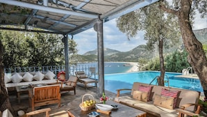 Área externa com mobiliário de madeira, piscina e vista para o mar e montanhas.