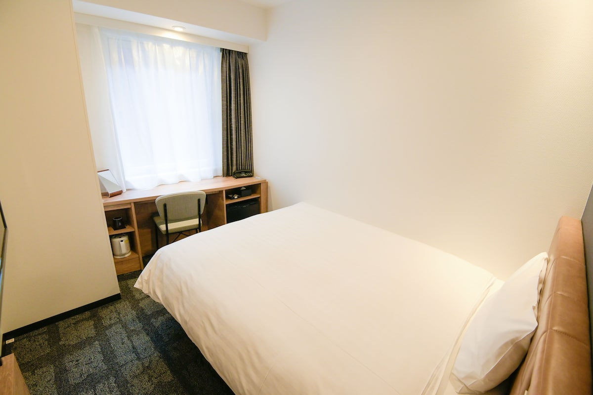 Et hotelværelse med seng, skrivebord med stol, et lille bord og et vindue med gardiner.