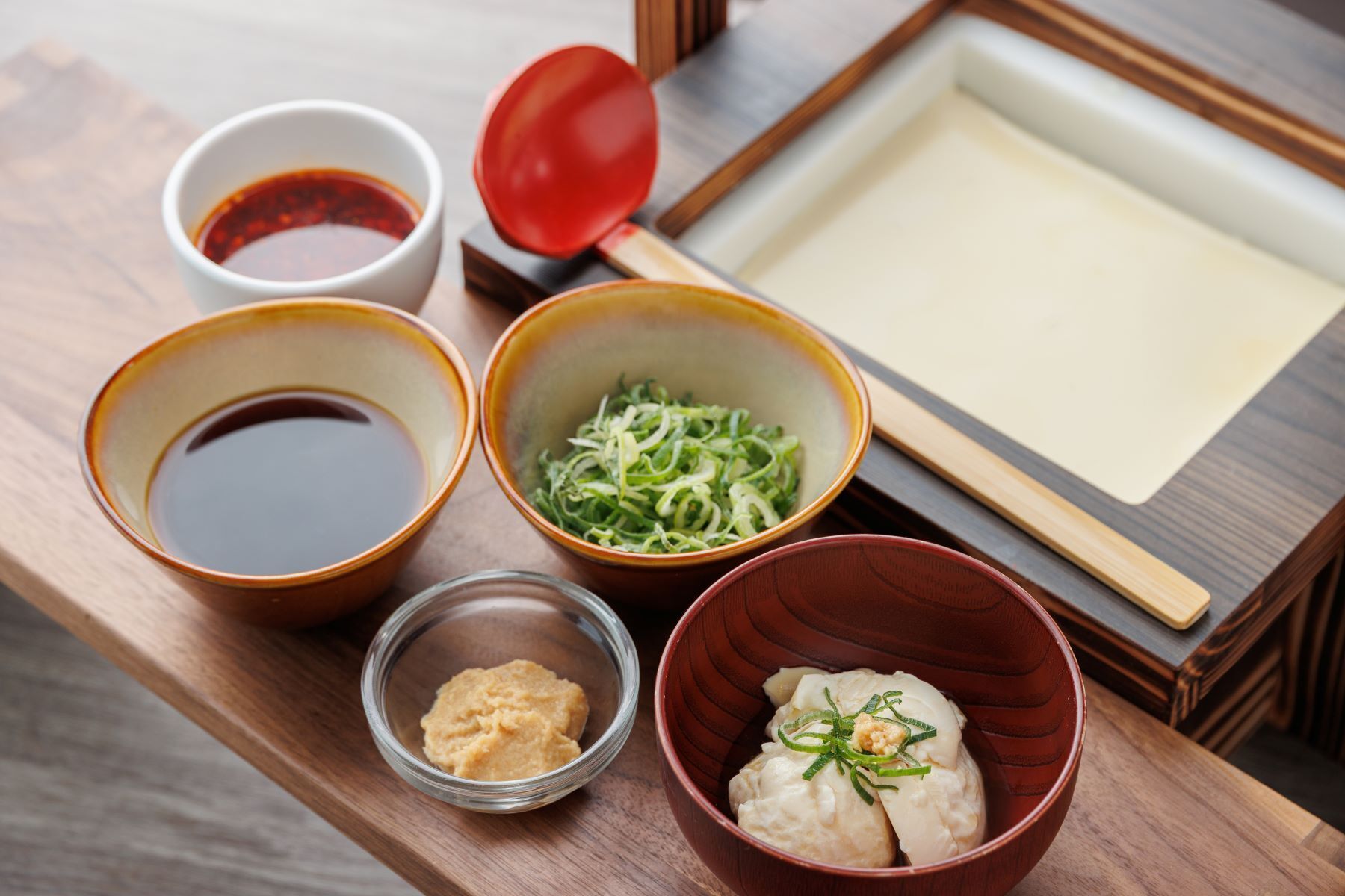 A traditional Japanese meal setup with bowls of soup, pickled vegetables, and dumplings, along with a small bowl of sauce and a red bowl with a lid.