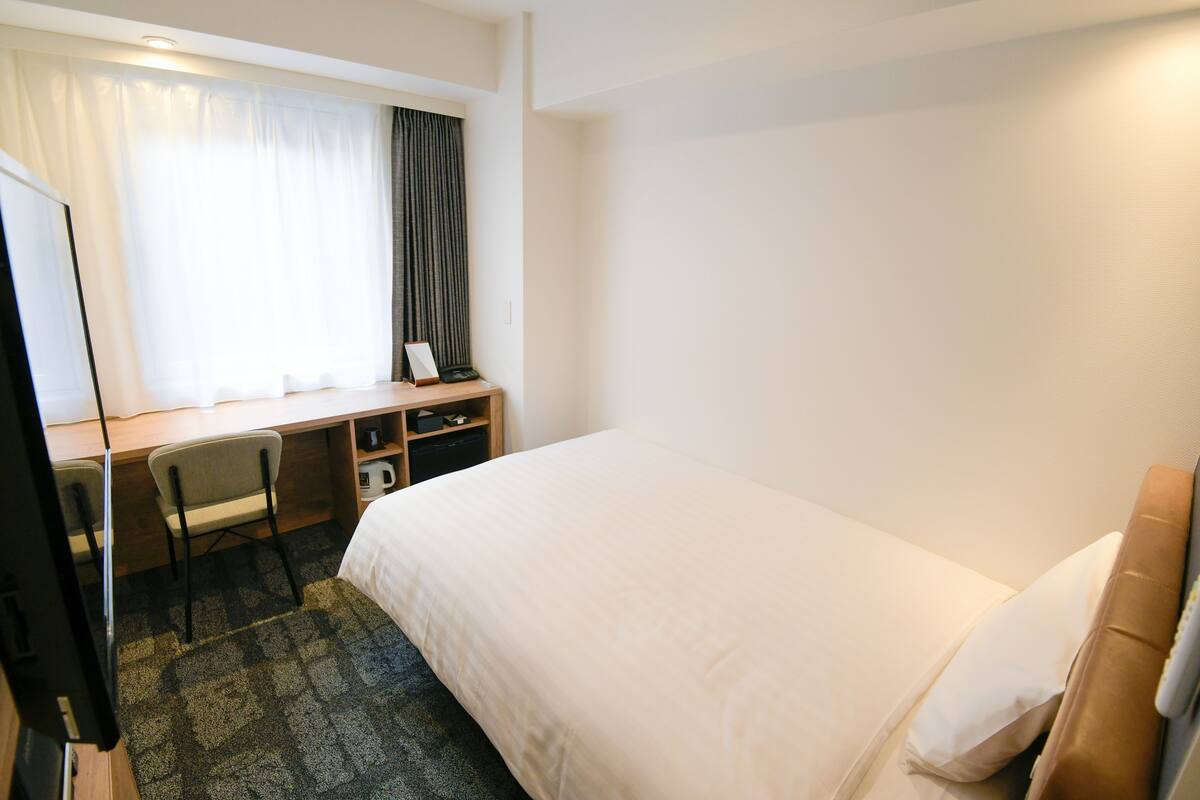 A hotel room with a bed, a desk with a chair, a television, and a window with curtains.